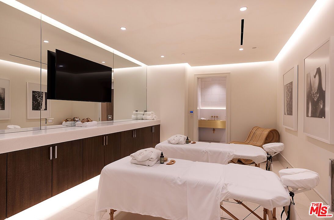 This image depicts a professional-style spa or massage room featuring two treatment tables draped in white linens. The room is designed with a minimalist aesthetic, incorporating dark wood cabinetry, a large wall-mounted television, and soft recessed lighting that creates a serene atmosphere. A curved wooden lounge chair sits in the corner, and a doorway leads to an adjacent room with a gold-toned sink area.