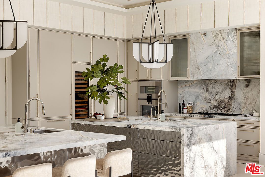 This sophisticated kitchen features a large central island with a stone countertop and a textured metallic base, complemented by a secondary sink area. The space is defined by light-toned cabinetry, a striking stone backsplash, and two modern pendant lights hanging above the island. A large potted plant serves as a focal point on the island, adding a touch of organic warmth to the sleek, high-end design.