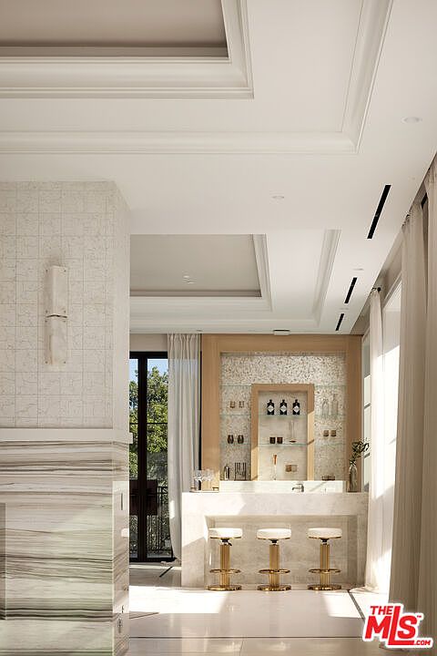 This elegant home bar area features a stone-clad counter with three gold-accented bar stools, set against a backdrop of light wood cabinetry and a decorative tiled wall. The space is illuminated by recessed lighting and a wall-mounted sconce, creating a sophisticated atmosphere. A large window with sheer curtains allows natural light to filter in, while the foreground showcases a textured stone pillar.
