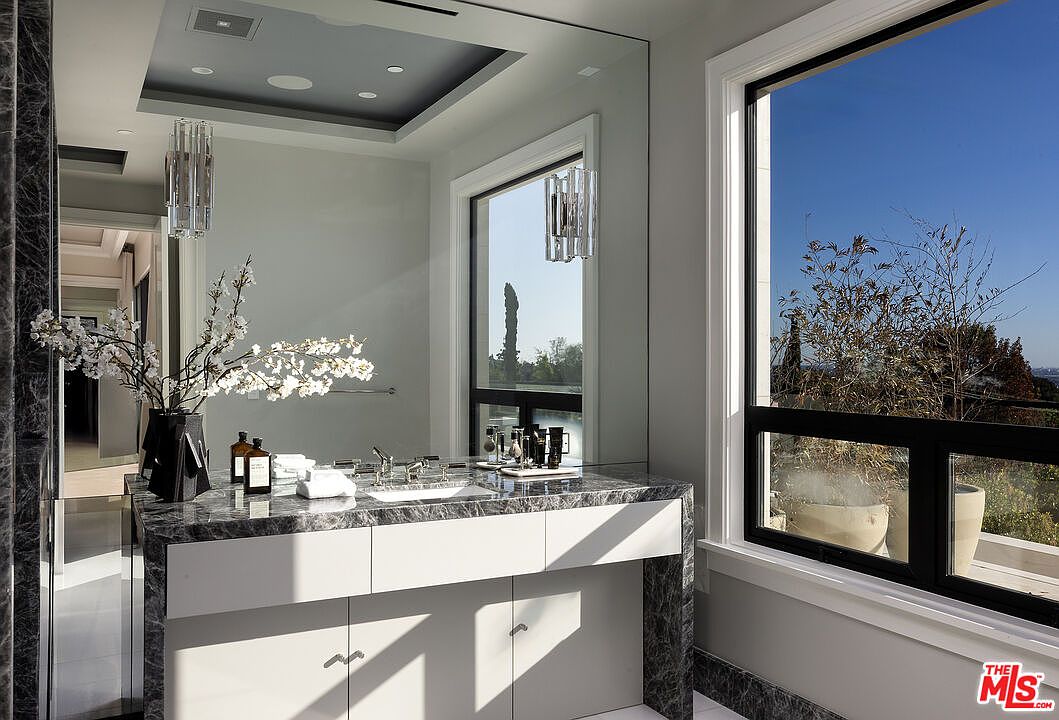 This elegant primary bathroom features a vanity topped with dark, veined stone and a sleek, integrated sink. A large, wall-spanning mirror reflects the room's sophisticated lighting and a window that provides a bright, scenic view of the outdoors. The space is characterized by its clean lines, modern fixtures, and a refined, minimalist aesthetic.