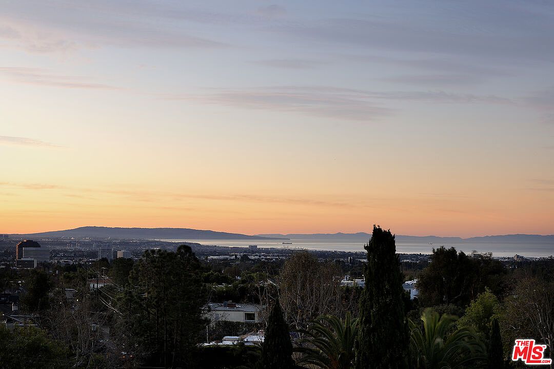 This image captures a sweeping, elevated panoramic view of a coastal landscape at sunset, showcasing a sprawling urban area meeting the ocean. In the foreground, lush treetops and palm fronds frame the scene, while the middle ground reveals a residential neighborhood leading toward the distant coastline and mountains. The sky is painted in soft hues of orange and blue, creating a serene and expansive atmosphere that highlights the property's desirable vantage point.