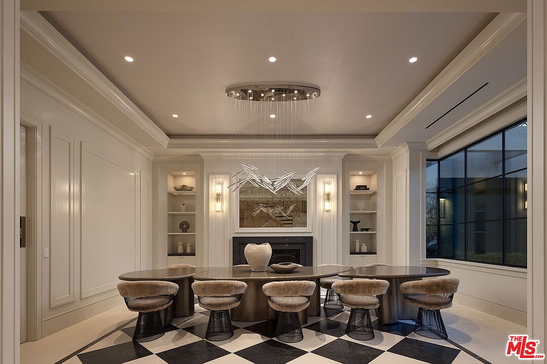 This sophisticated dining room features a large, dark-toned oval table surrounded by five modern, textured chairs with wire-frame bases. The space is anchored by a striking, multi-tiered chandelier suspended above the table, set against a backdrop of a dark fireplace surround and symmetrical built-in shelving. A bold black-and-white checkered floor pattern adds a dramatic flair to the room, which is illuminated by recessed lighting and wall sconces.