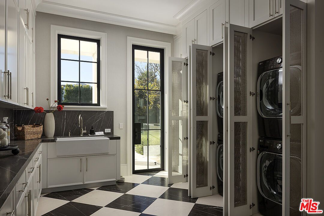 This sophisticated laundry room features a striking black-and-white checkered tile floor and custom white cabinetry paired with dark stone countertops. A farmhouse-style sink is positioned beneath a window, while a set of stacked laundry appliances is neatly concealed behind mesh-paneled doors. The space is bright and airy, with a glass-paned door providing a view of the outdoor greenery, creating a clean and functional aesthetic.