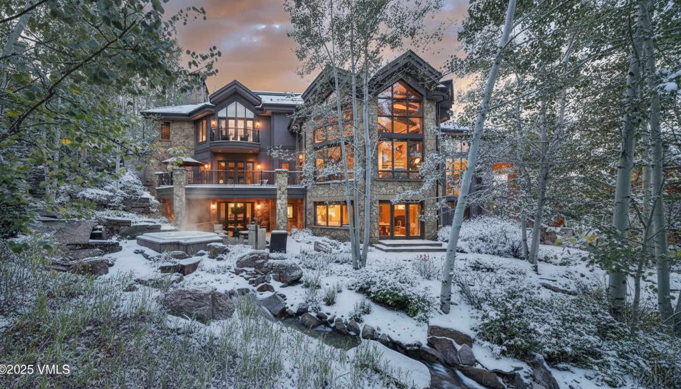 This image showcases the rear exterior of a luxurious home during winter. The architecture features stone and dark siding, large windows, and a multi-level deck with a hot tub. The landscape is snowy, with trees framing the house, contributing to a serene and picturesque setting.