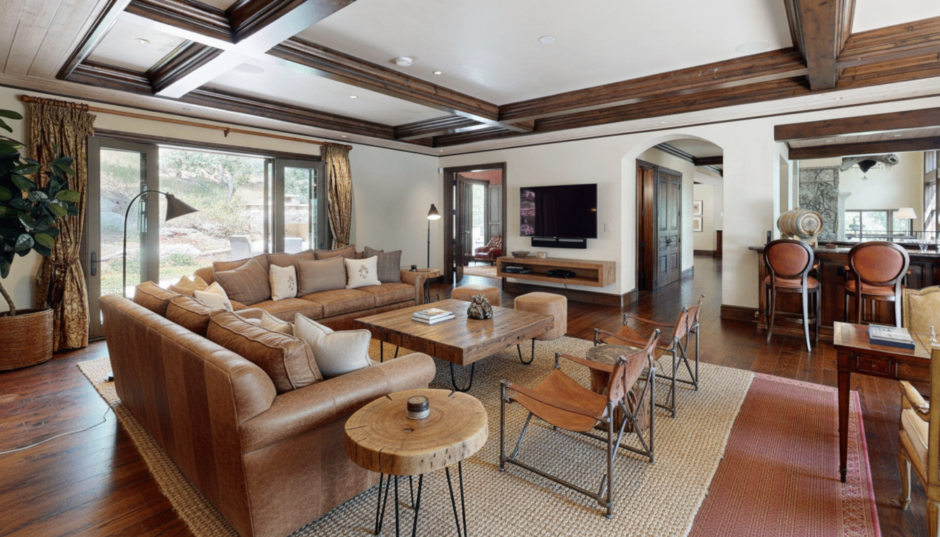 This is a warm and inviting living room featuring a comfortable leather sectional sofa, a rustic wooden coffee table, and stylish accent chairs. The room boasts exposed wooden beams on the ceiling and hardwood floors, contributing to a sophisticated yet cozy atmosphere. Large windows provide ample natural light and a view of the outdoors.