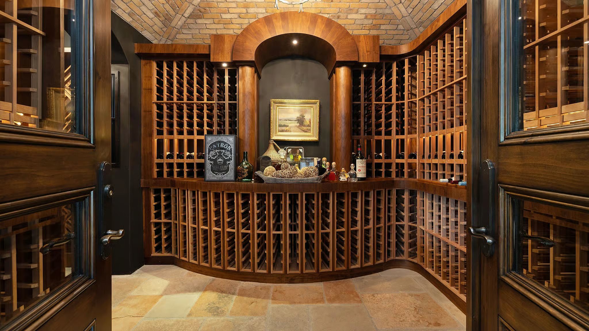 This is a well-appointed wine cellar featuring custom wooden racking designed to store numerous bottles of wine. The cellar has a curved design and includes display space with a picture and other decorative items. The room is accessed by wood framed glass doors.