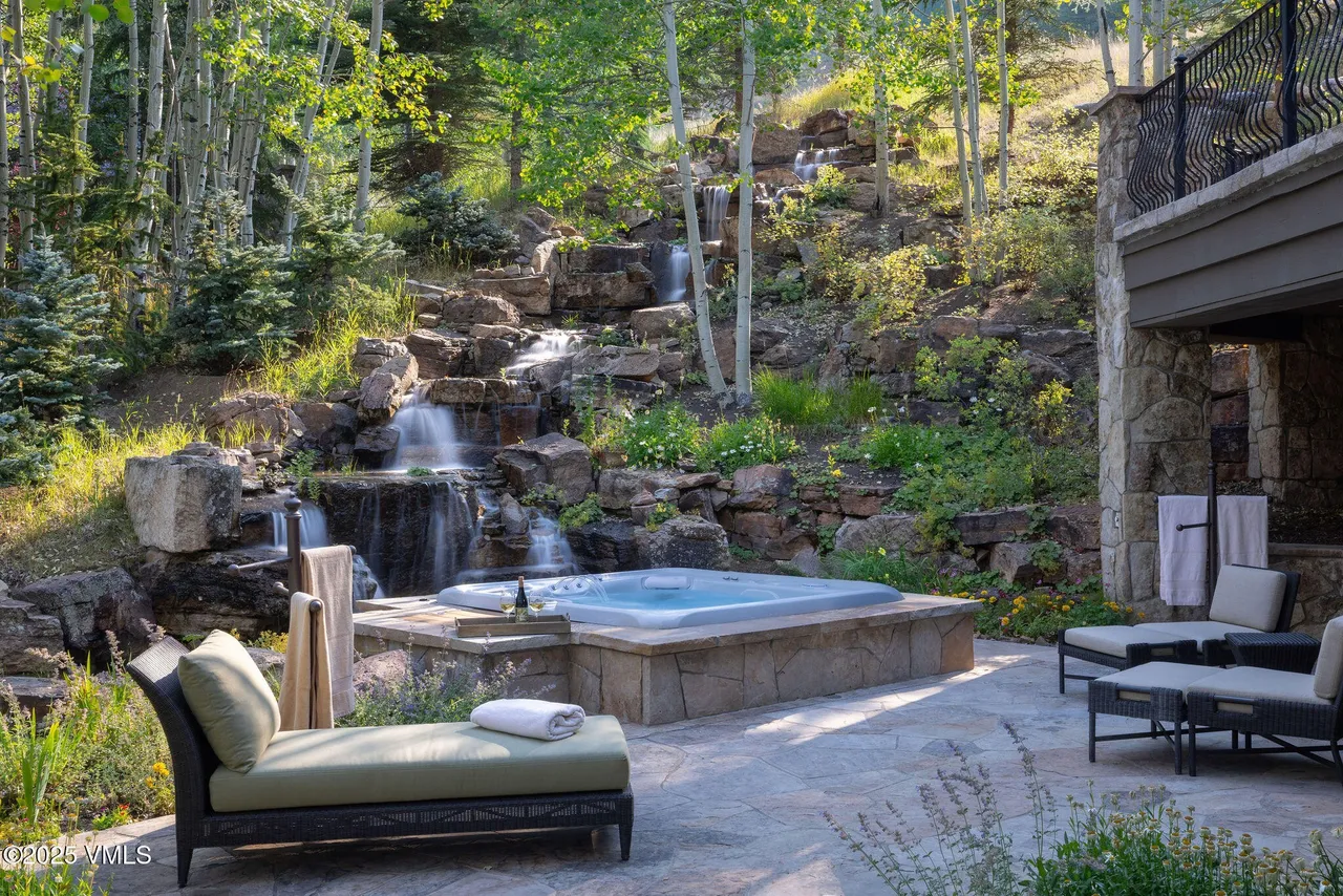 This exterior shot showcases a luxurious patio area complete with a built-in hot tub set against a stunning waterfall backdrop. The space features stone paving, comfortable lounge seating, and a tranquil ambiance, enhancing the property's appeal for relaxation and entertainment. Part of the home's balcony is visible on the right.