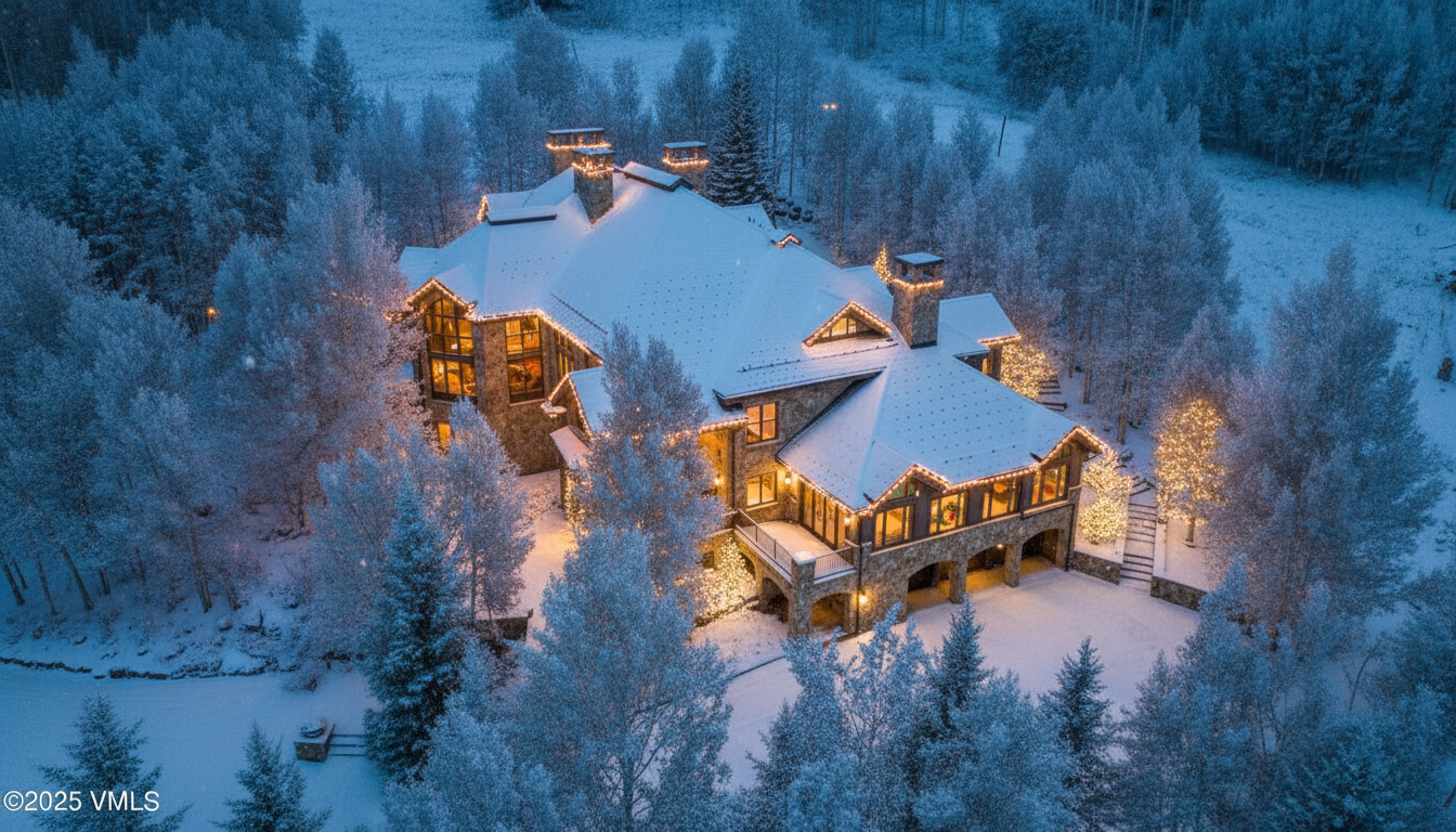 This aerial view captures a stunning, snow-covered luxury home, blending beautifully with the surrounding forest. The house is adorned with festive lights, highlighting its architectural details and creating a warm, inviting ambiance. The landscape is pristine and wintry, emphasizing the privacy and serenity of the property.