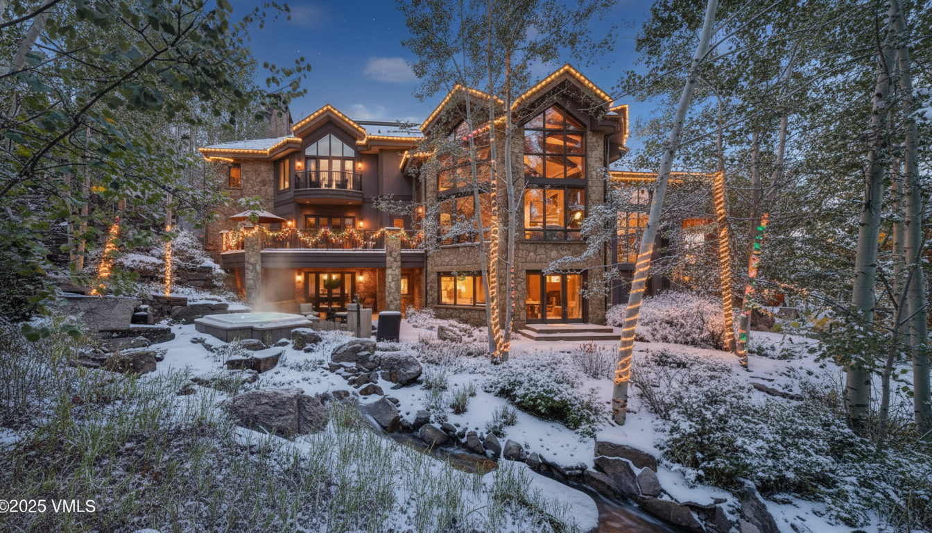 This image shows the rear exterior of a luxury home in a snowy setting. The house features multiple stories, a stone facade, and large windows providing ample natural light. A hot tub is visible in the foreground, suggesting a relaxing outdoor space complemented by the surrounding trees and elegant lighting that gives the property a warm and inviting glow.