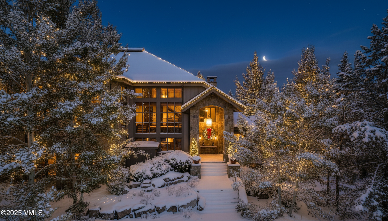 This is a picturesque front view of a home in a winter setting, likely intended for a real estate listing. The house is decorated with holiday lights, and a wreath adorns the entrance, creating a warm and inviting ambiance. Snow-covered trees and shrubs surround the property, highlighting the serene and luxurious atmosphere of the location.