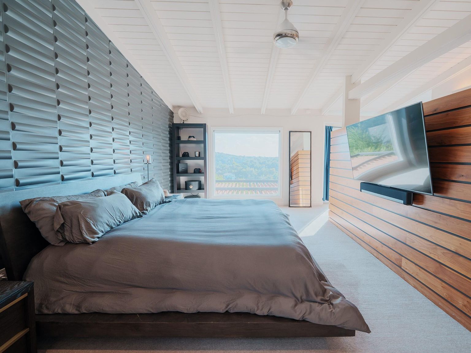 This is a primary bedroom featuring a modern design with a textured gray accent wall behind the bed. The room has a large bed with gray linens, a wooden platform frame, and a built-in shelving unit. A flat-screen TV is mounted on a wooden paneled wall, and a window provides natural light and a view of the outdoors.