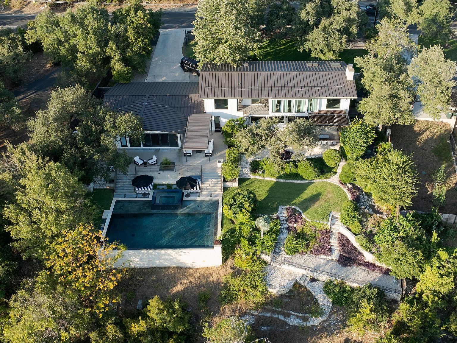 This aerial shot showcases a luxurious property featuring a modern house with a dark metal roof, a swimming pool, and a spa. The landscape is meticulously designed with lush greenery, stone pathways, and manicured lawns, creating a serene and private oasis. The property is surrounded by mature trees, enhancing its secluded feel.