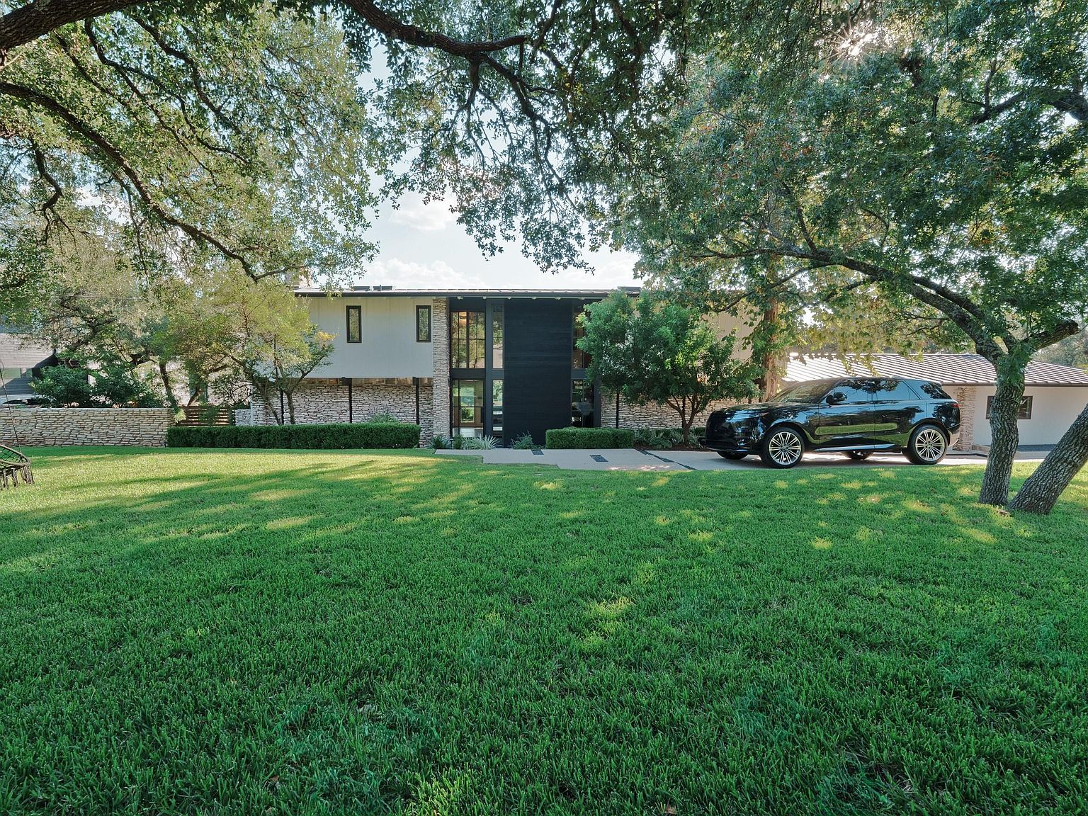 This is a front view of a modern house with a well-manicured lawn. The house features a combination of stone and siding, with a dark-colored central section that includes the entryway. A black SUV is parked in the driveway, and mature trees frame the house, adding to its curb appeal.