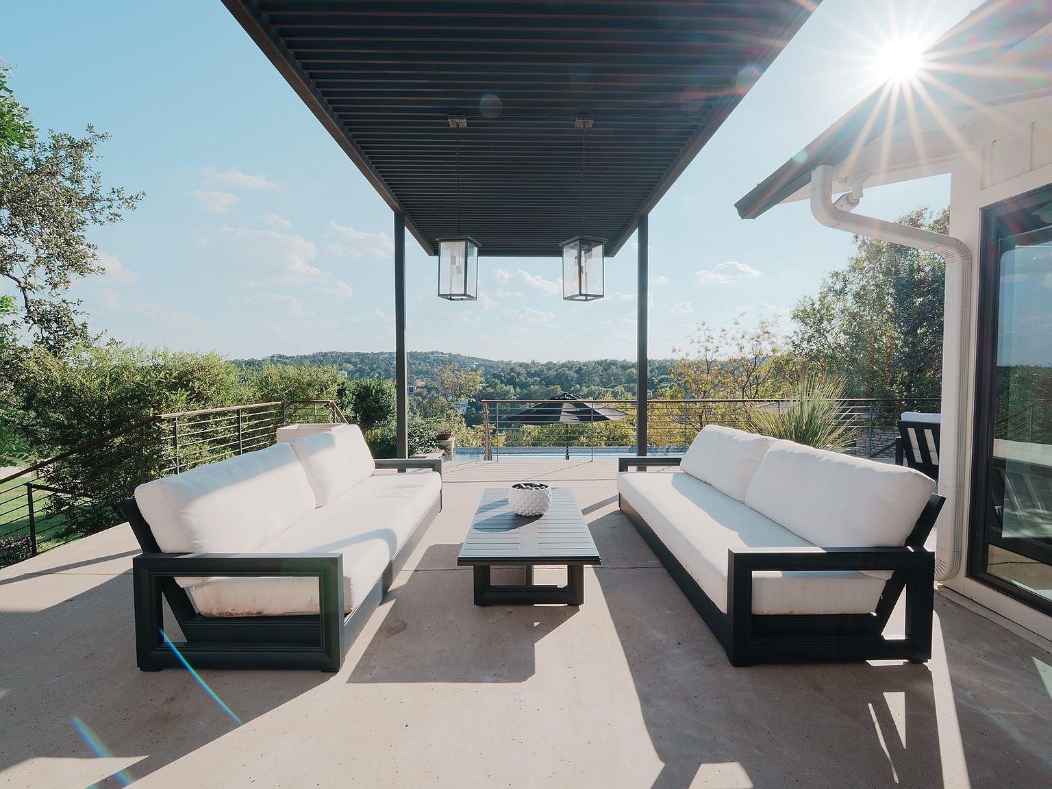 This image showcases a stylish outdoor patio or deck area, featuring two modern sofas with white cushions and dark frames, arranged around a sleek coffee table. An overhead pergola provides shade, while pendant lights add an elegant touch. The space offers a beautiful view of the surrounding landscape, creating a relaxing and inviting atmosphere.