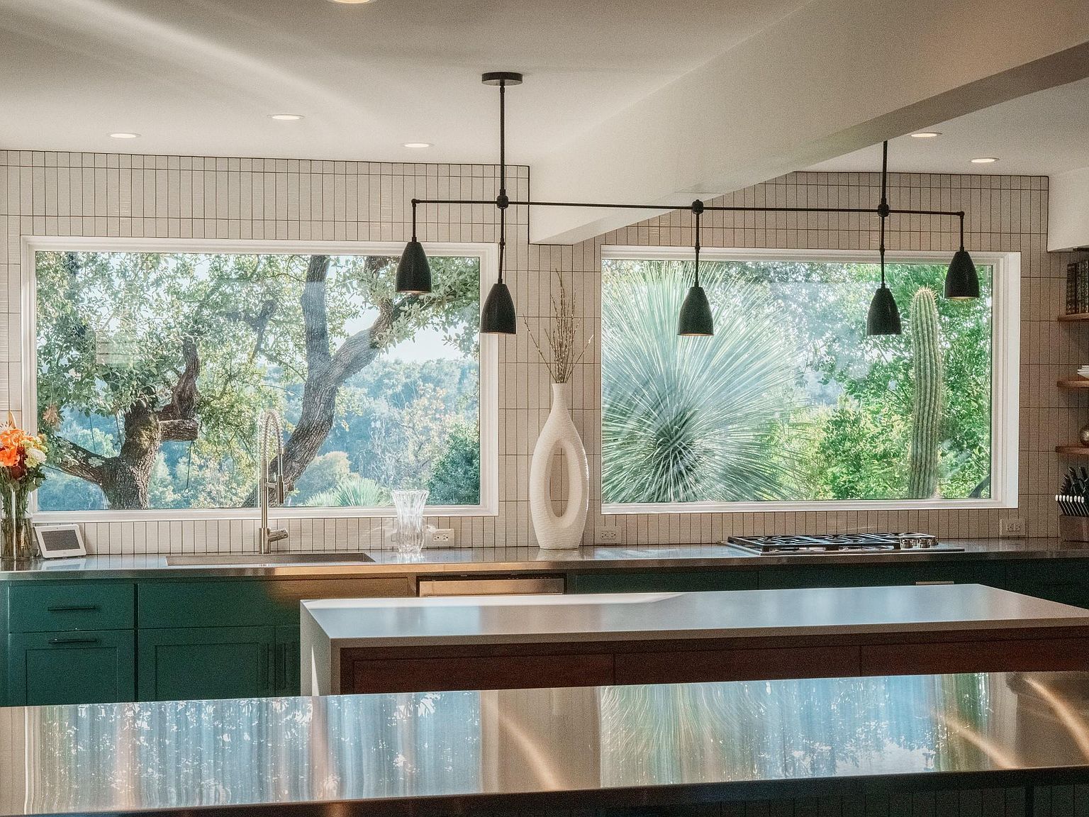 This is a bright and modern kitchen featuring a large window with a view of lush greenery. The kitchen has dark green cabinetry, stainless steel countertops, and a central island with a light-colored countertop. The lighting fixtures are modern pendant lights, and the overall style is contemporary and inviting.