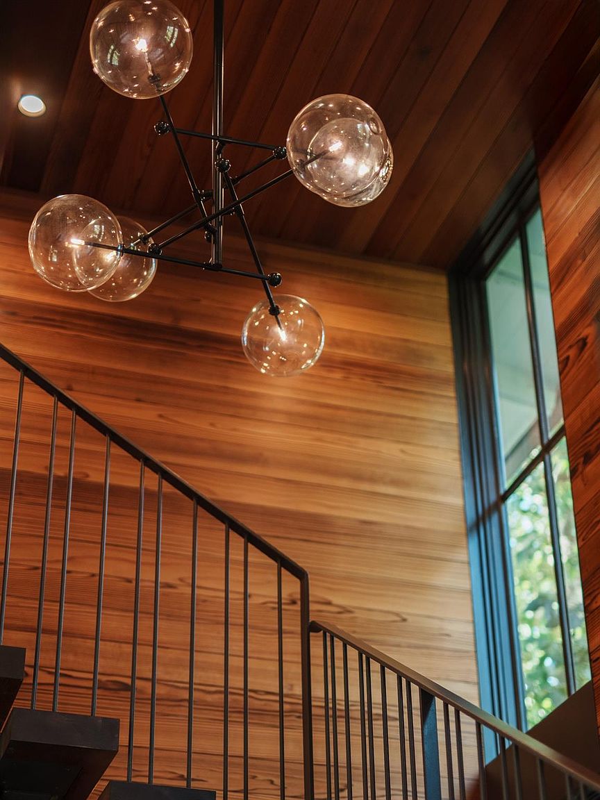 This interior shot showcases a modern staircase with dark metal railings and wooden steps. The walls are clad in warm-toned wood paneling, complemented by a unique chandelier featuring multiple glass globes. A large window provides natural light, enhancing the architectural details and creating an inviting atmosphere.