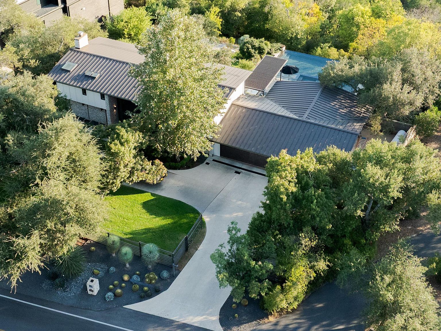 This aerial shot showcases a modern home nestled among lush greenery. The house features a striking metal roof, a well-manicured lawn, and a contemporary driveway design. The surrounding trees provide privacy and enhance the property's natural appeal, making it a secluded and desirable residence.