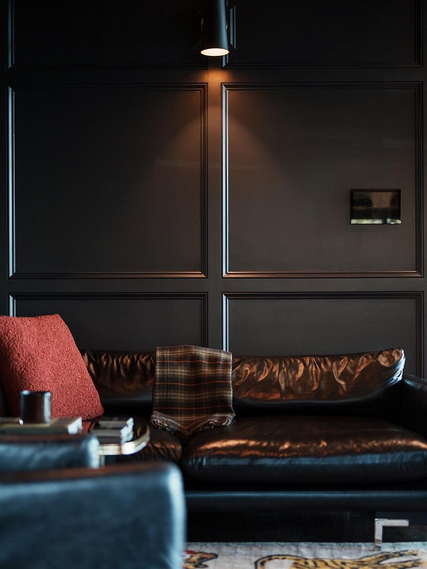 This image showcases a sophisticated living room with dark, paneled walls and a luxurious leather sofa adorned with a plaid throw and a red pillow. A sleek, modern light fixture illuminates the scene, highlighting the room's elegant and moody ambiance. The foreground features a glimpse of another seating area, suggesting a spacious and well-appointed interior.