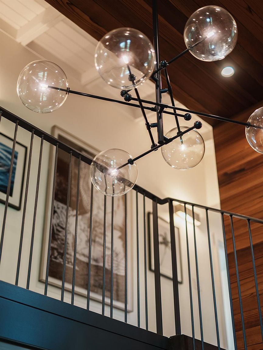 This interior shot showcases a modern staircase with black metal railings and a unique, multi-bulb chandelier. Artwork adorns the walls, adding a touch of sophistication to the space. The warm wood ceiling contrasts with the white walls, creating a visually appealing and inviting atmosphere.