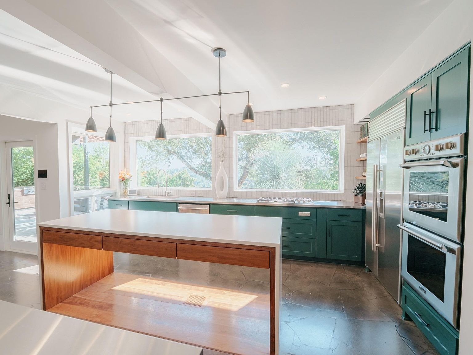 This is a bright and spacious kitchen featuring dark green cabinetry, stainless steel appliances, and a large island with a white countertop and wood base. Large windows provide ample natural light and views of the outdoors. The flooring is a polished concrete, adding to the modern aesthetic.
