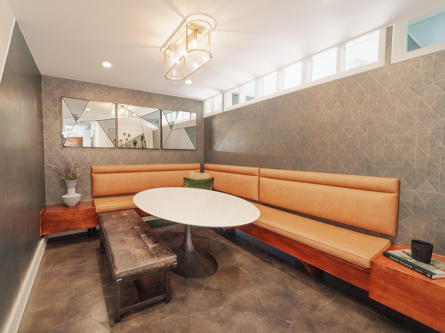 This dining area features a built-in banquette with tan leather upholstery, complemented by a round white table and a dark leather bench. The room is decorated with geometric mirrors and a modern light fixture, creating a stylish and inviting space. The flooring is polished concrete, adding an industrial touch.