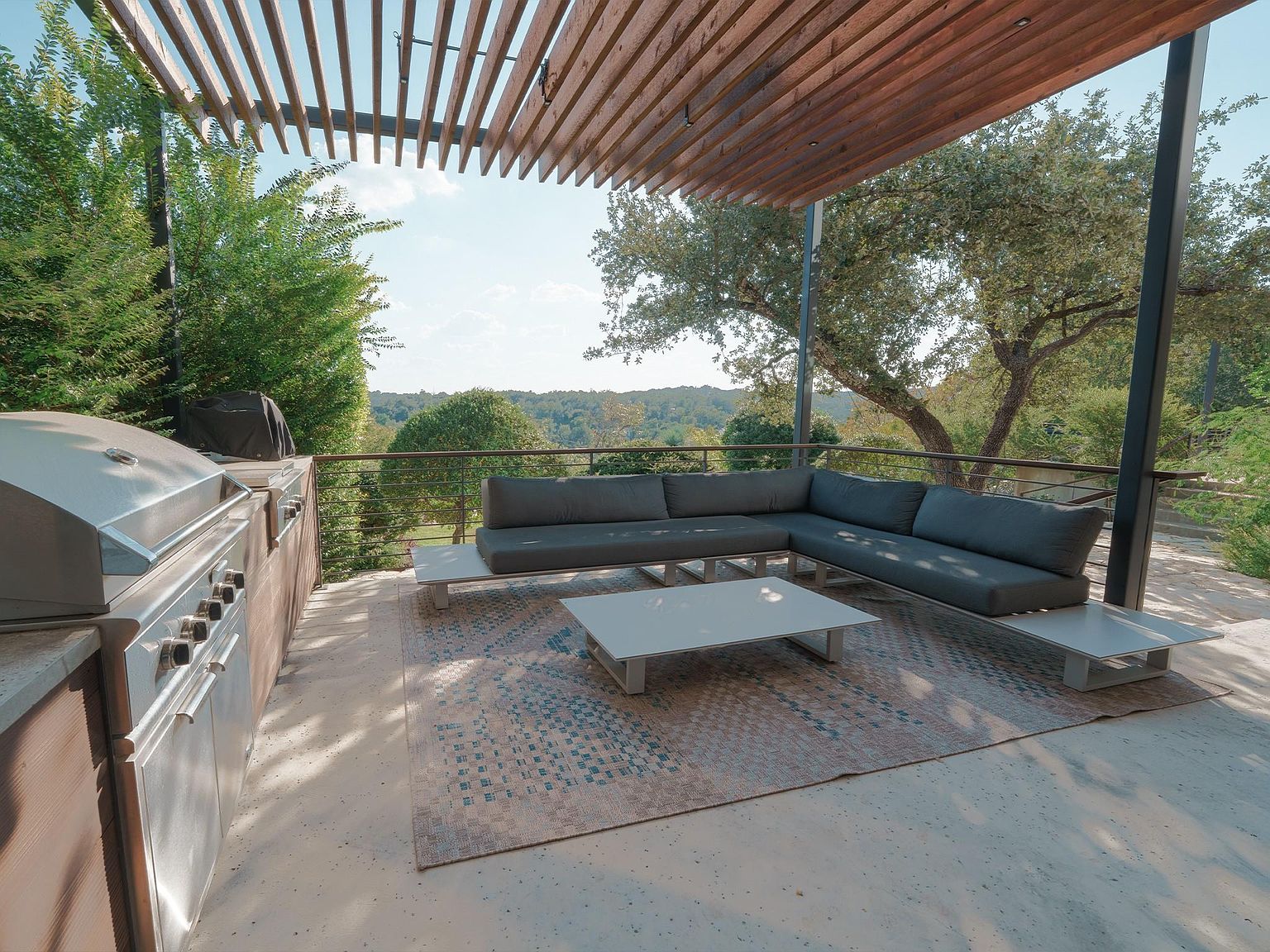 This image showcases a stylish outdoor patio area, perfect for entertaining or relaxing. The space features a comfortable sectional sofa, a modern coffee table, and a built-in grill area with stainless steel appliances. A wooden pergola provides shade, while the surrounding greenery and scenic view create a serene atmosphere.