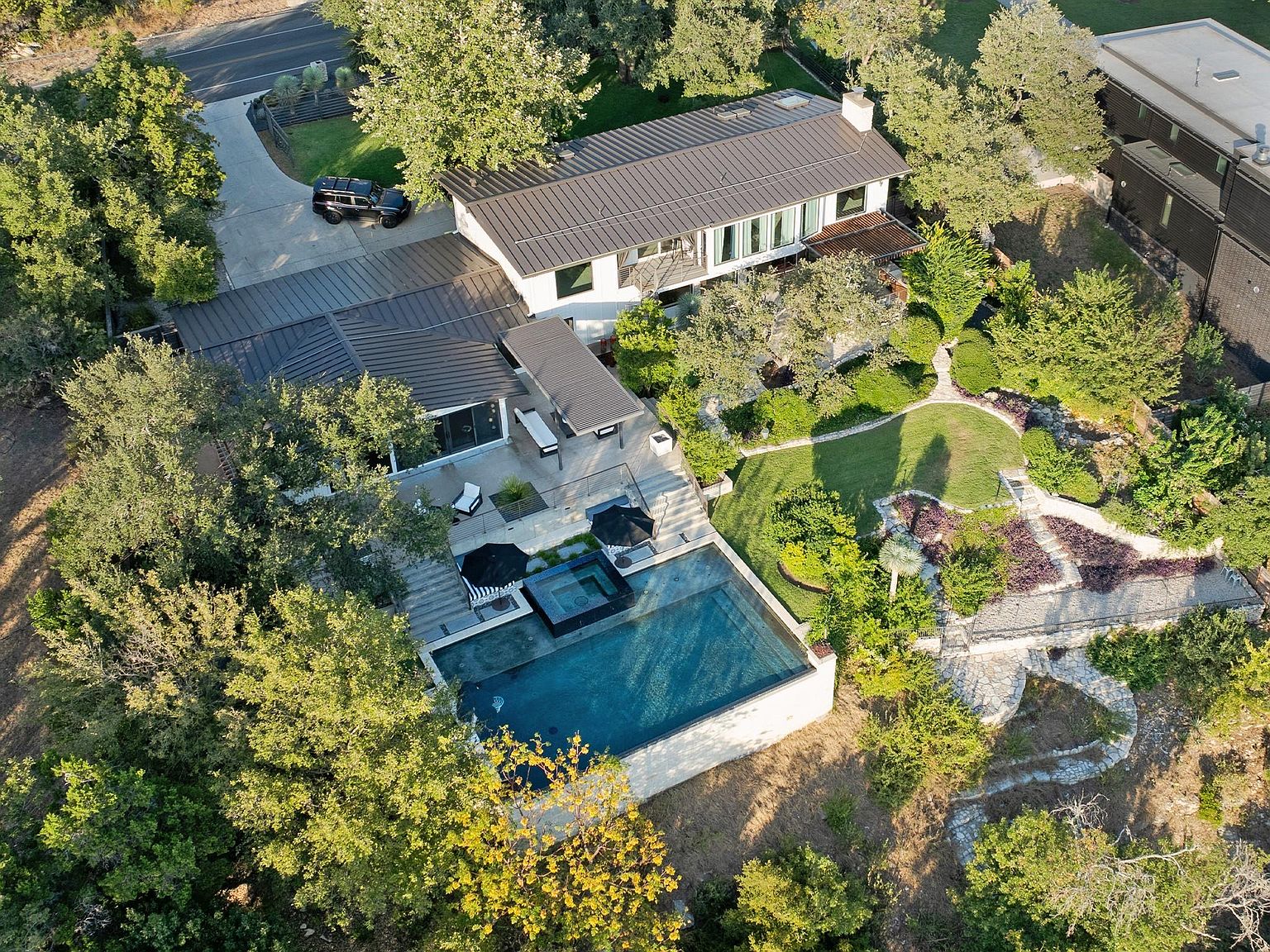 This aerial shot showcases a luxurious modern home with a dark metal roof, a pristine pool and spa area, and meticulously landscaped grounds. The property features a long driveway leading to the house, surrounded by mature trees and lush greenery, creating a private and serene atmosphere. The architectural design emphasizes clean lines and a seamless integration with the natural environment.