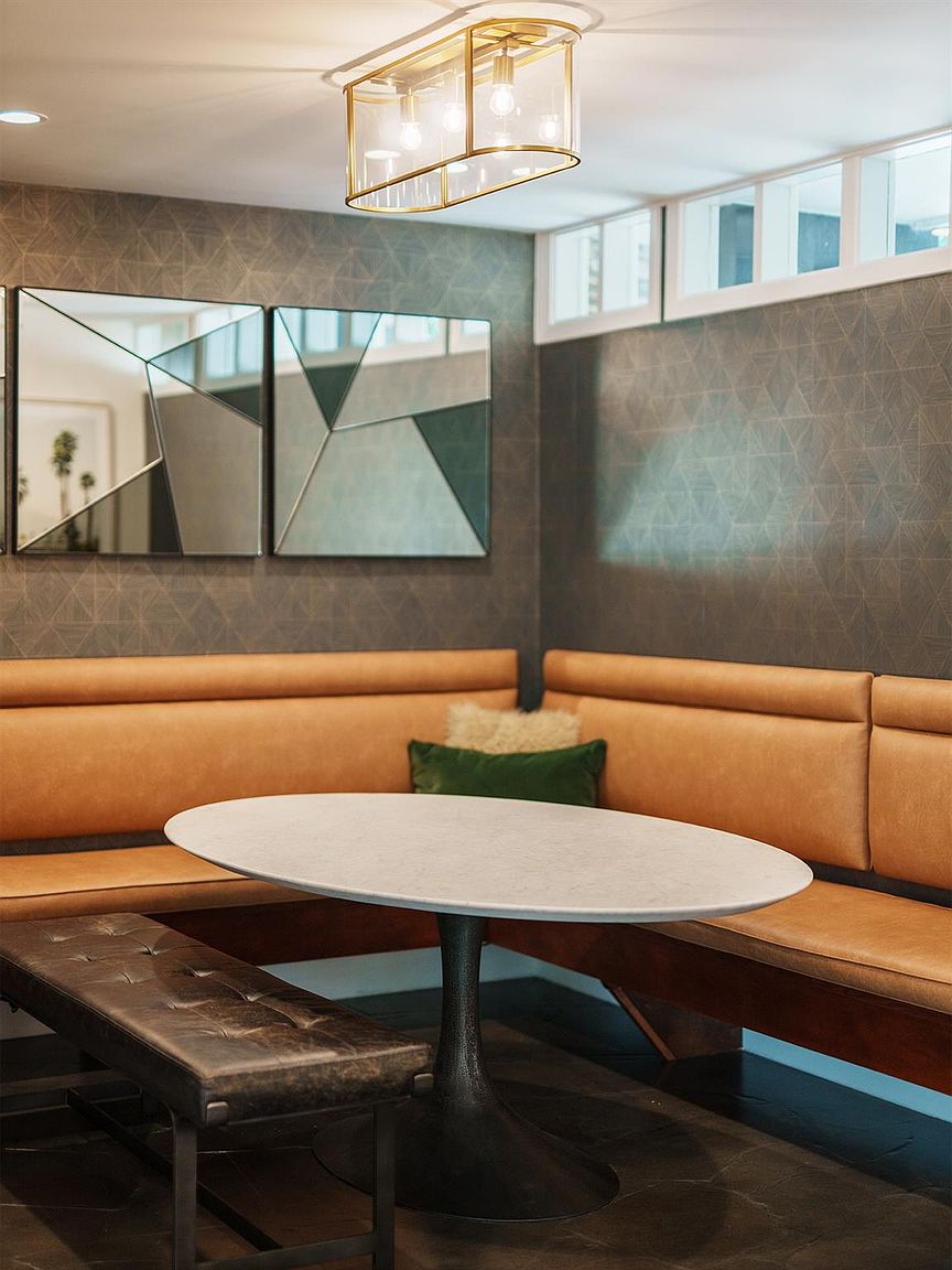 This interior shot showcases a cozy dining area with a built-in booth featuring tan leather upholstery and a dark leather bench. A white oval table sits in the center, illuminated by a modern, rectangular chandelier. The walls are adorned with geometric mirrors and patterned wallpaper, creating a stylish and inviting atmosphere.