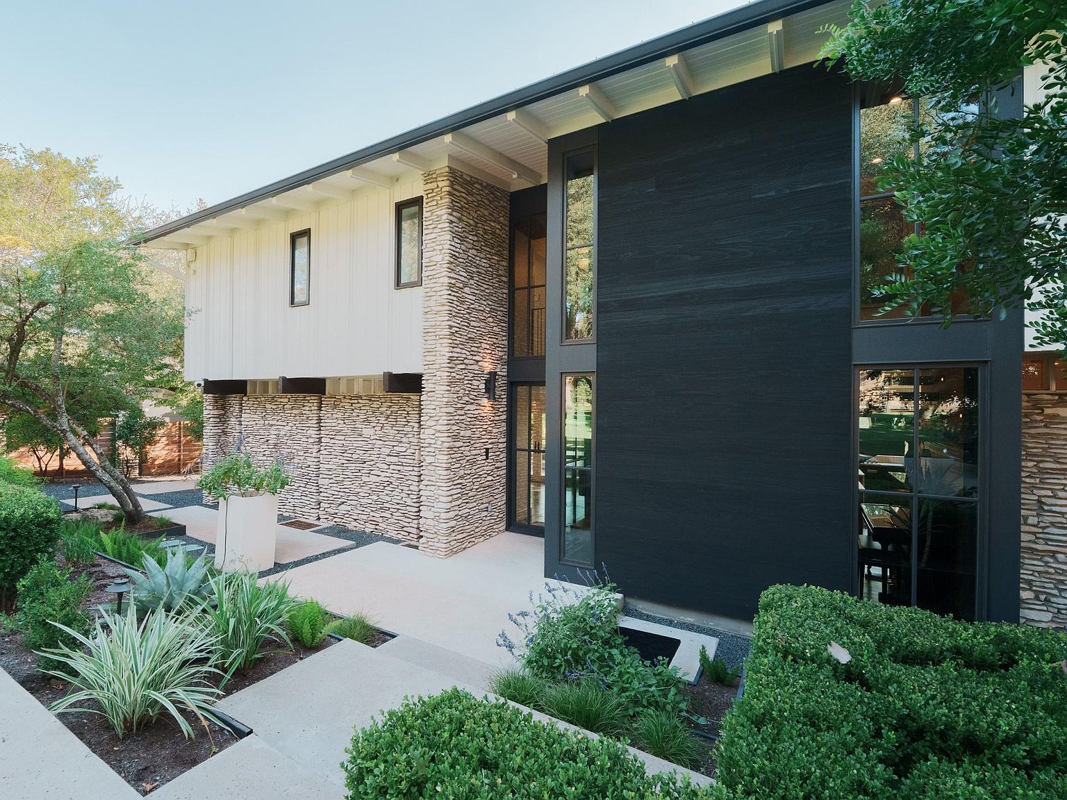 This is a striking front view of a modern home featuring a unique blend of materials. The facade incorporates dark wood paneling, light-colored stone accents, and white vertical siding. Large windows provide ample natural light, and the landscaping is meticulously designed with a mix of greenery and hardscaping, creating an inviting and sophisticated curb appeal.