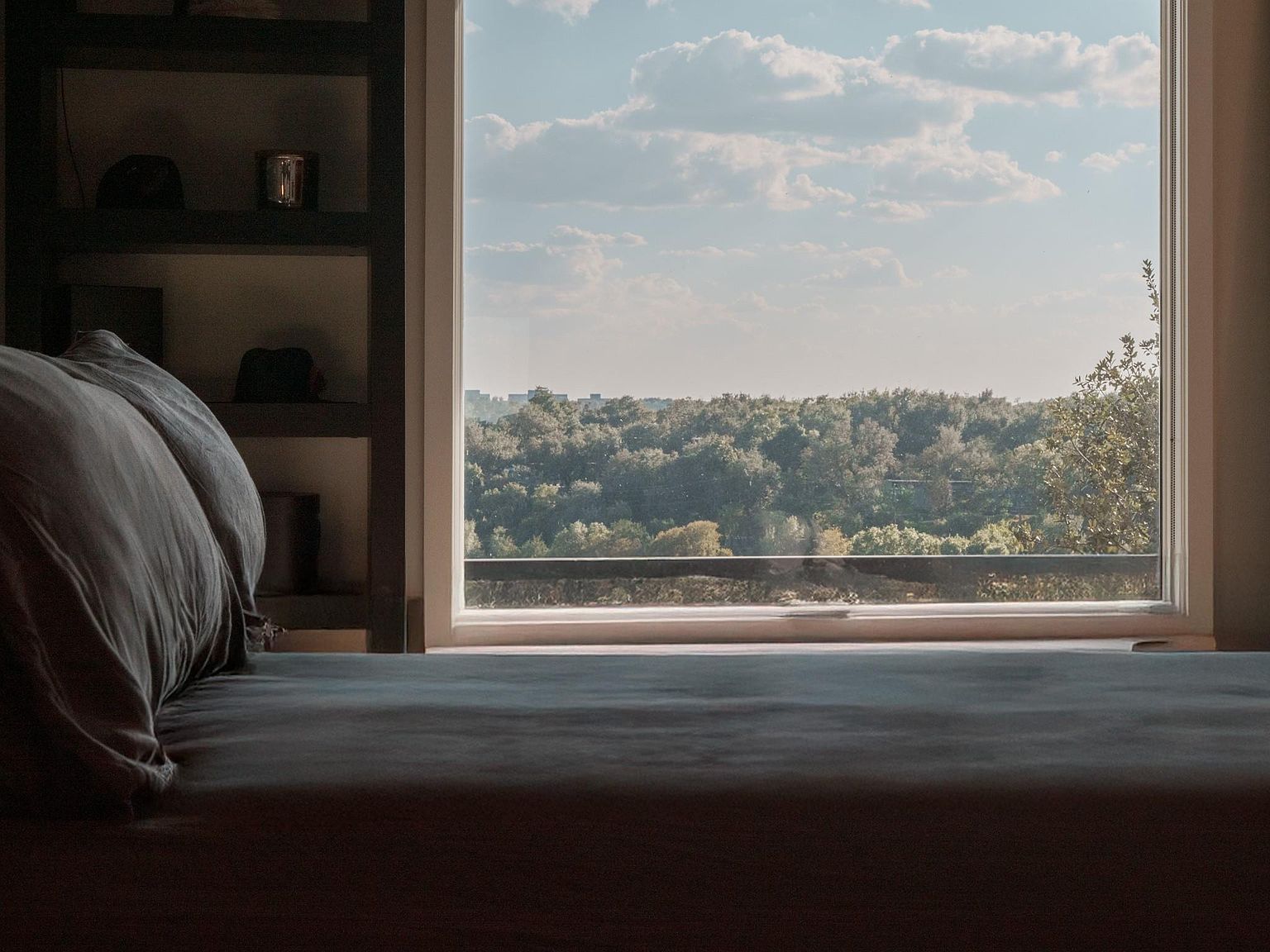 This image showcases a bedroom interior with a focus on a large window offering a scenic view of a lush, green landscape under a partly cloudy sky. The bed is prominently featured in the foreground, suggesting a comfortable and inviting space. A built-in bookshelf adds a touch of sophistication and storage to the room.