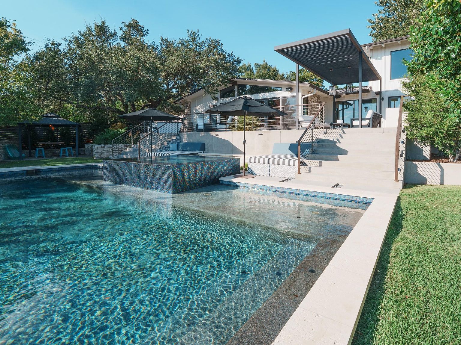 This image showcases a luxurious backyard pool and spa area. The pool features clear blue water and a mosaic tile accent wall, while the spa is elevated and surrounded by lounge seating. A modern pergola and well-manicured lawn add to the upscale ambiance, creating an inviting outdoor living space.