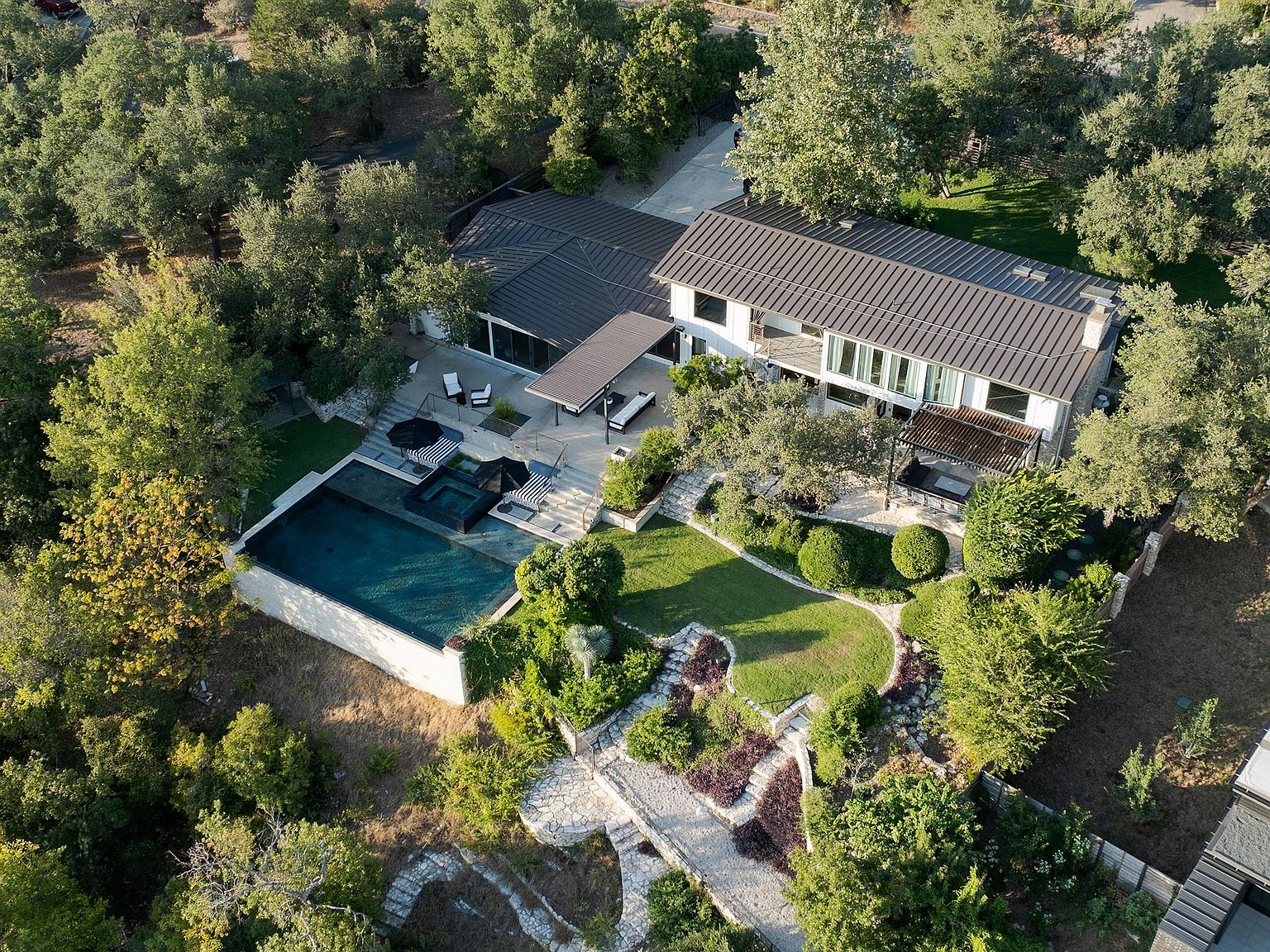 This aerial shot showcases a luxurious property featuring a modern house with a dark metal roof, a pristine pool and spa area, and meticulously landscaped grounds. The house is surrounded by lush greenery, providing a sense of privacy and tranquility. Stone pathways and tiered gardens add to the property's appeal, creating a serene and inviting outdoor space.
