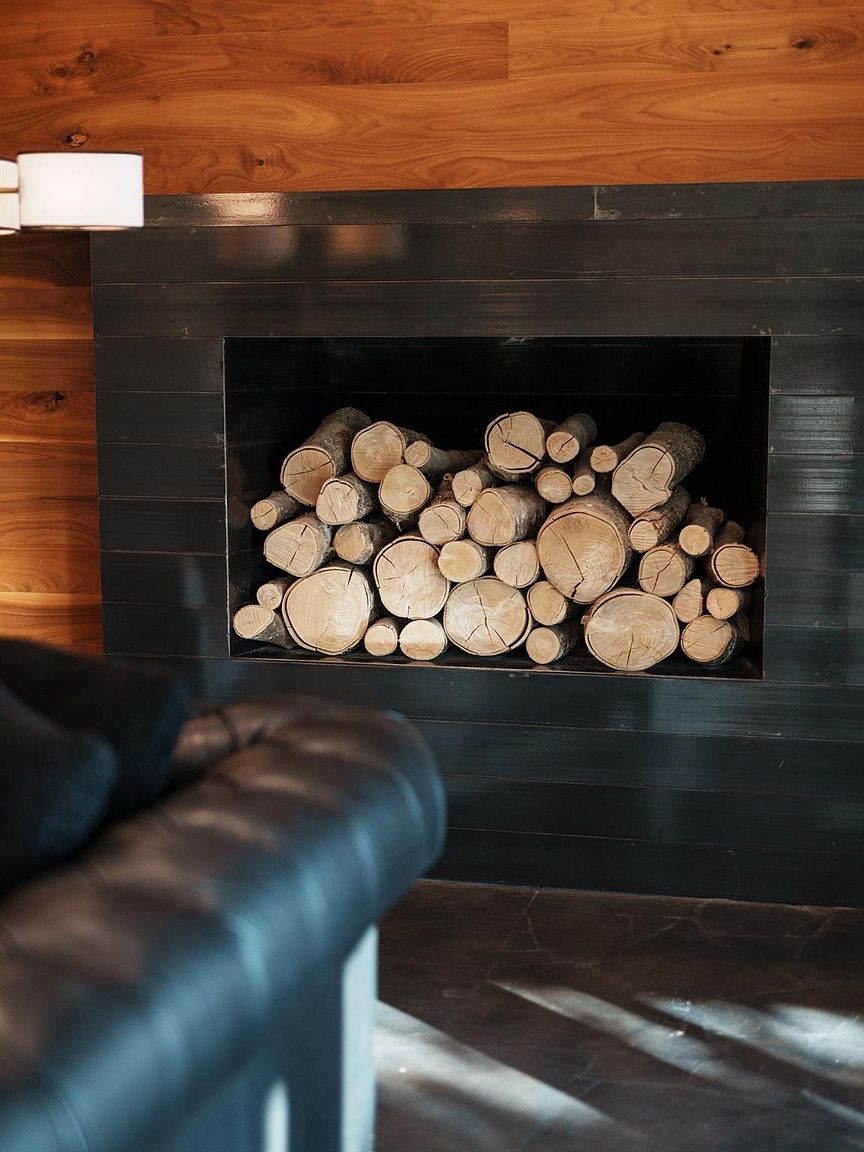 The image showcases a modern living room detail, focusing on a built-in fireplace filled with neatly stacked logs. The fireplace surround is a dark, horizontally-paneled material, contrasting with the warm wood paneling on the adjacent wall. A portion of a dark leather sofa is visible in the foreground, adding depth and suggesting a comfortable seating area.