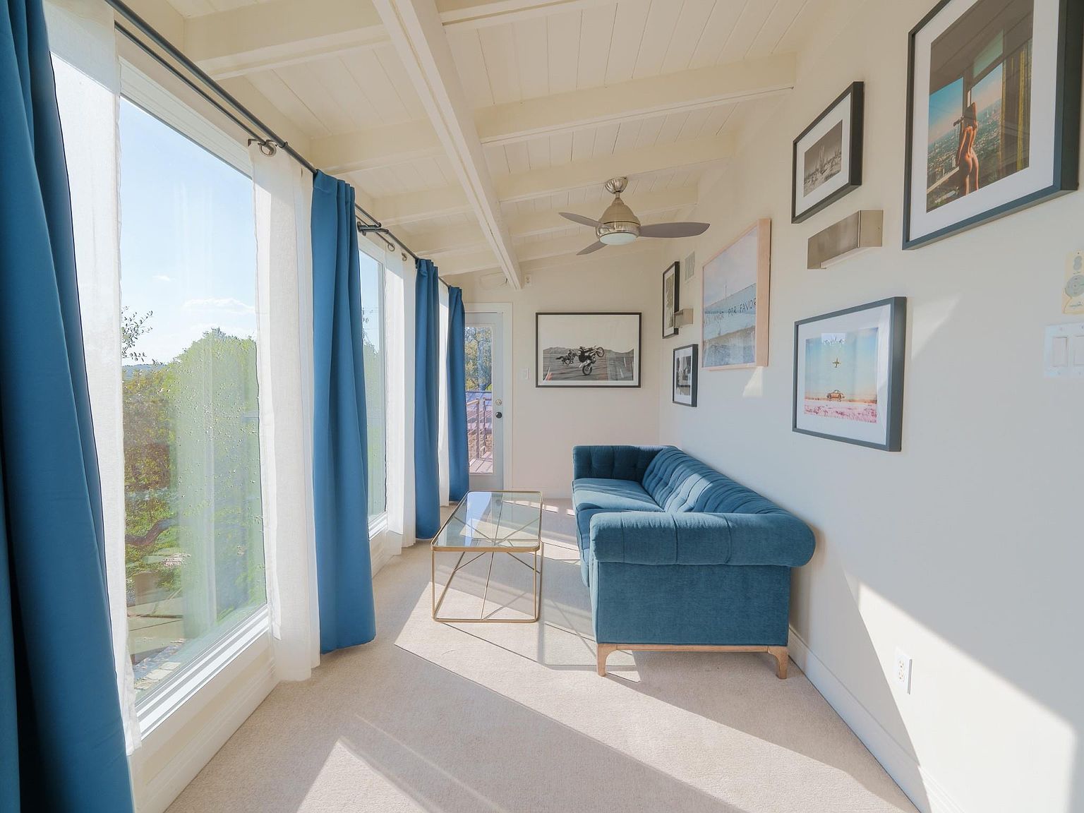 This is an interior shot of a sun-drenched living room featuring a comfortable blue sofa, a glass coffee table, and a wall adorned with framed artwork. Large windows with blue and white curtains offer a view of the outdoors, while the white ceiling with exposed beams adds a touch of architectural interest. The room exudes a bright and airy atmosphere, perfect for relaxation and entertaining.