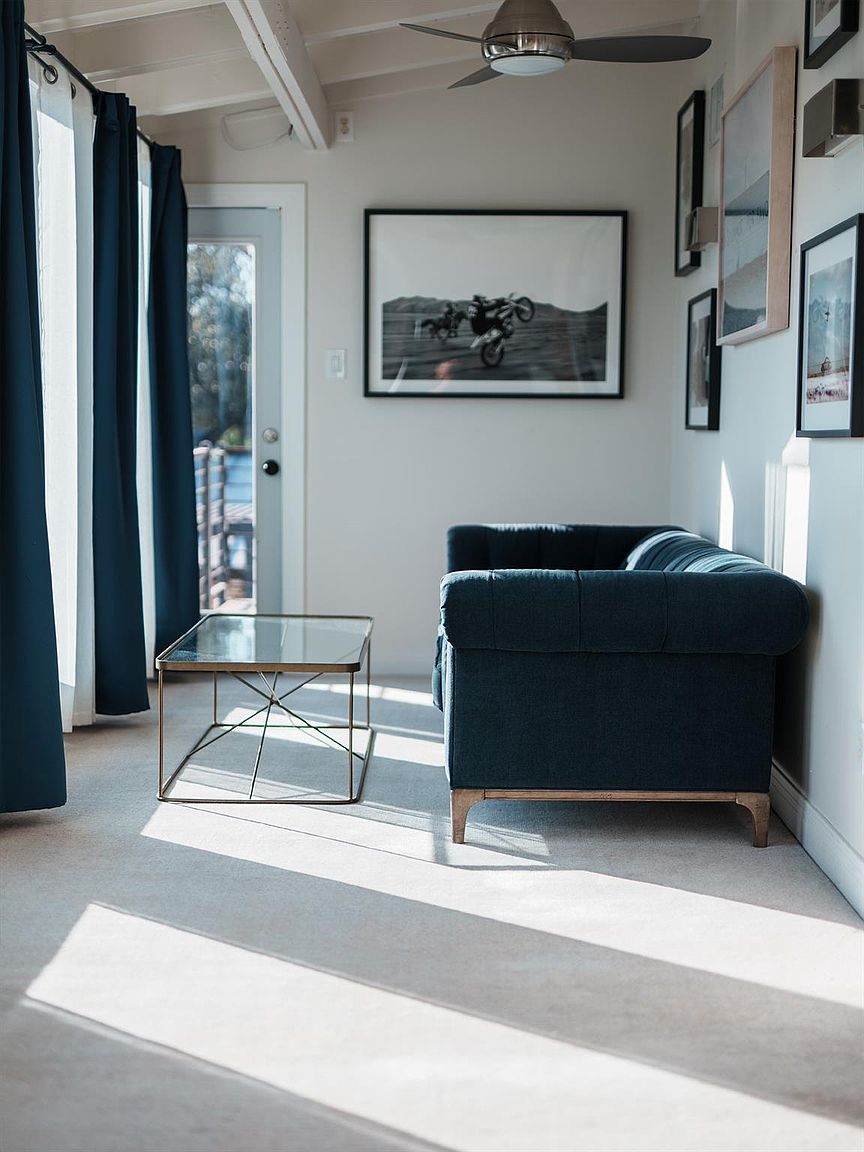 This is an interior shot of a living room featuring a blue sofa, a glass-topped coffee table with a metal frame, and dark blue curtains. Artwork adorns the walls, and natural light streams in through a door, creating a bright and airy atmosphere. The room has a modern and minimalist aesthetic.