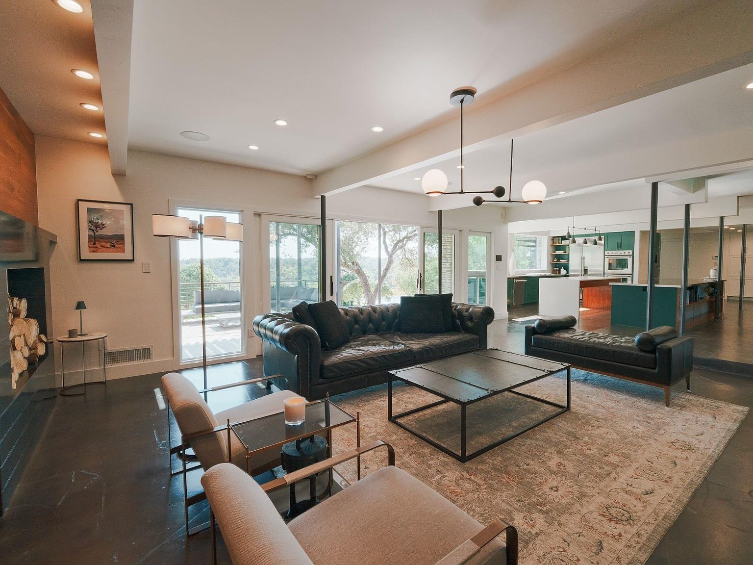 This is a spacious living room featuring a dark leather sofa and loveseat, a large area rug, and modern lighting fixtures. The room has an open floor plan that flows into the kitchen, with large windows providing natural light and views of the outdoors. The overall style is contemporary and luxurious, creating a comfortable and inviting atmosphere.