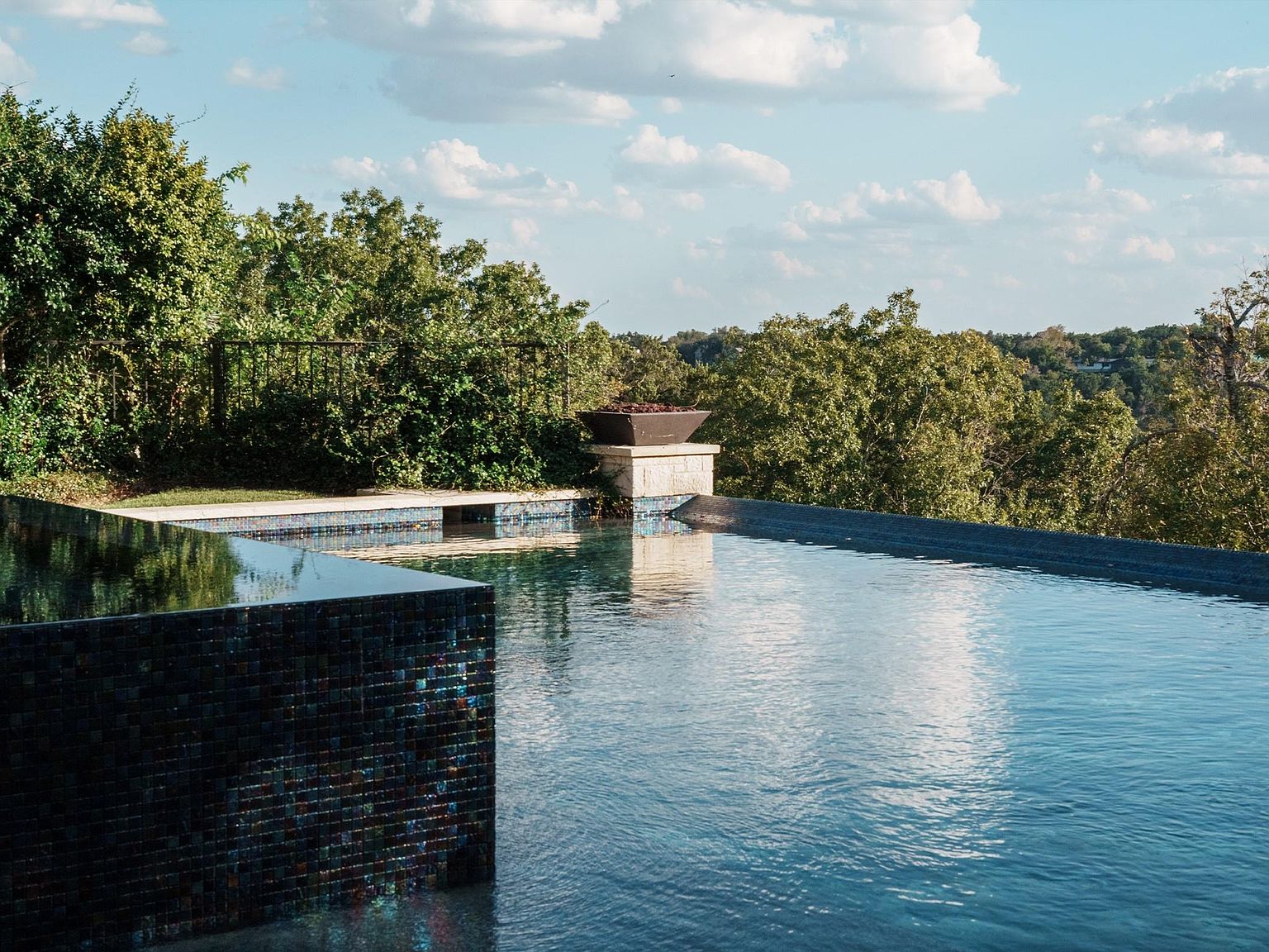 This image showcases a luxurious infinity pool with dark blue tiling, reflecting the sky and surrounding trees. The pool's edge blends seamlessly with the landscape, offering a sense of tranquility and privacy. A modern fire feature adds a touch of elegance and warmth to the outdoor space.