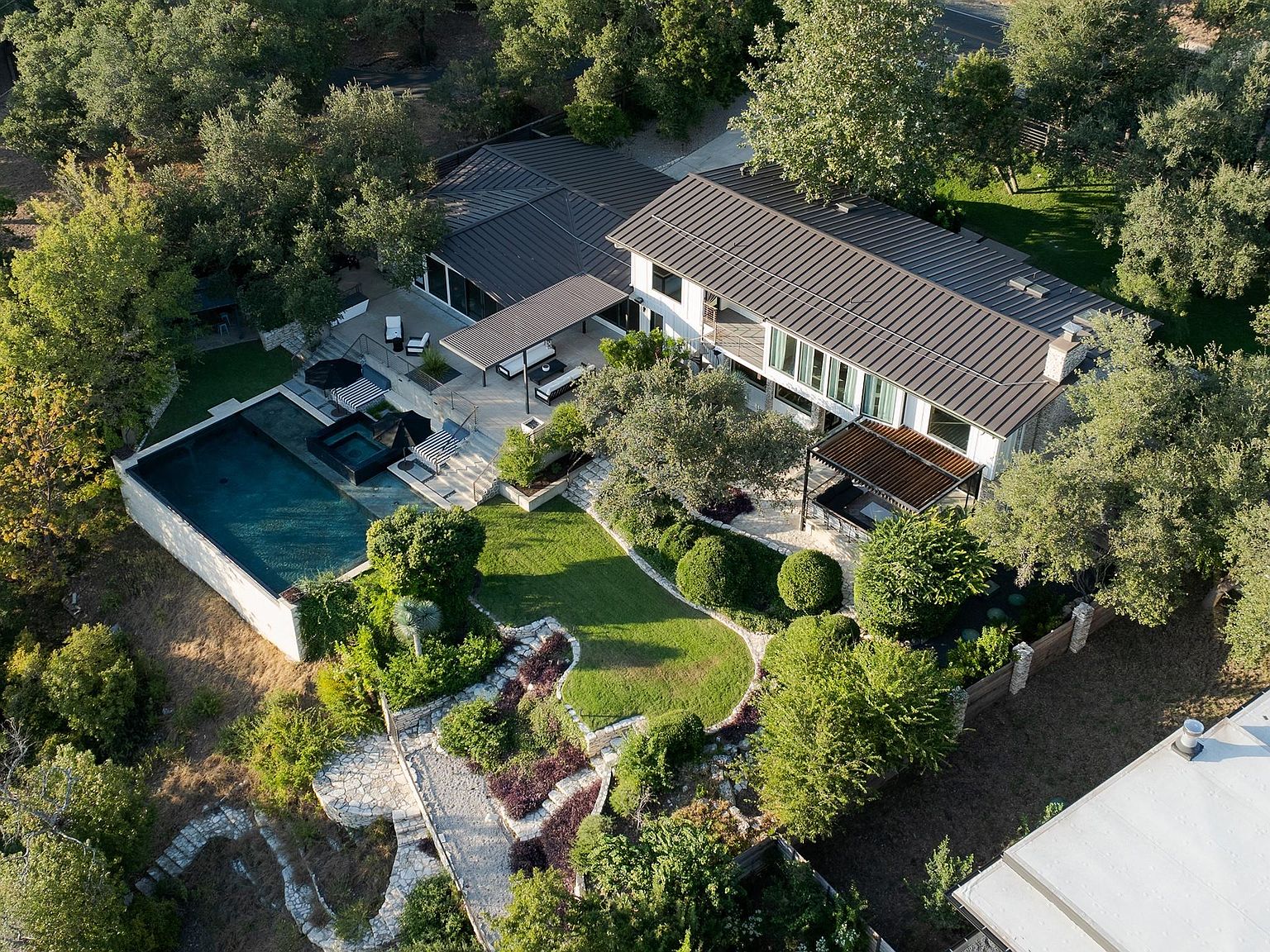 This aerial view showcases a luxurious modern home nestled among lush greenery. The property features a dark-roofed house with a light-colored exterior, a stunning pool and spa area, and meticulously landscaped gardens with stone pathways. The overall impression is one of privacy, elegance, and high-end living.