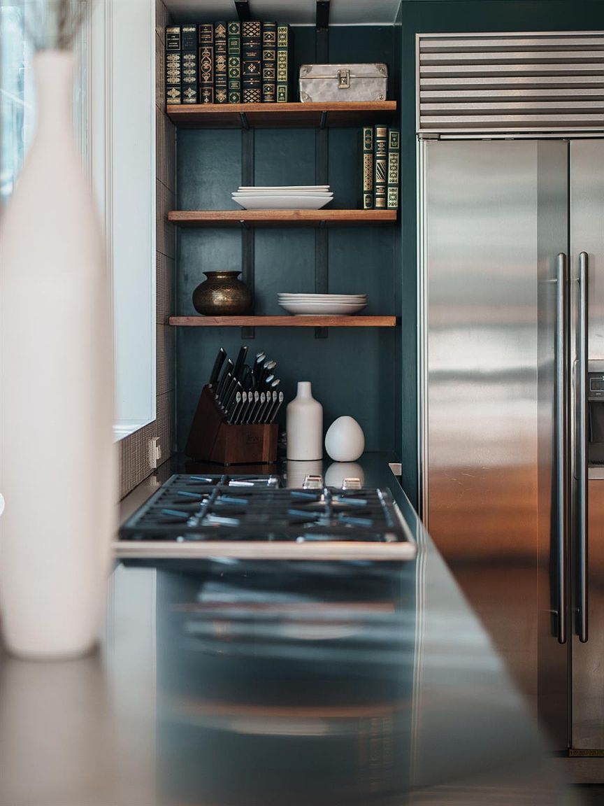 This is a detailed shot of a modern kitchen featuring a stainless steel refrigerator and a gas stovetop. The dark teal cabinetry is complemented by wooden shelves displaying books and dishware. The countertop is a sleek, reflective surface, adding to the kitchen's sophisticated and functional design.