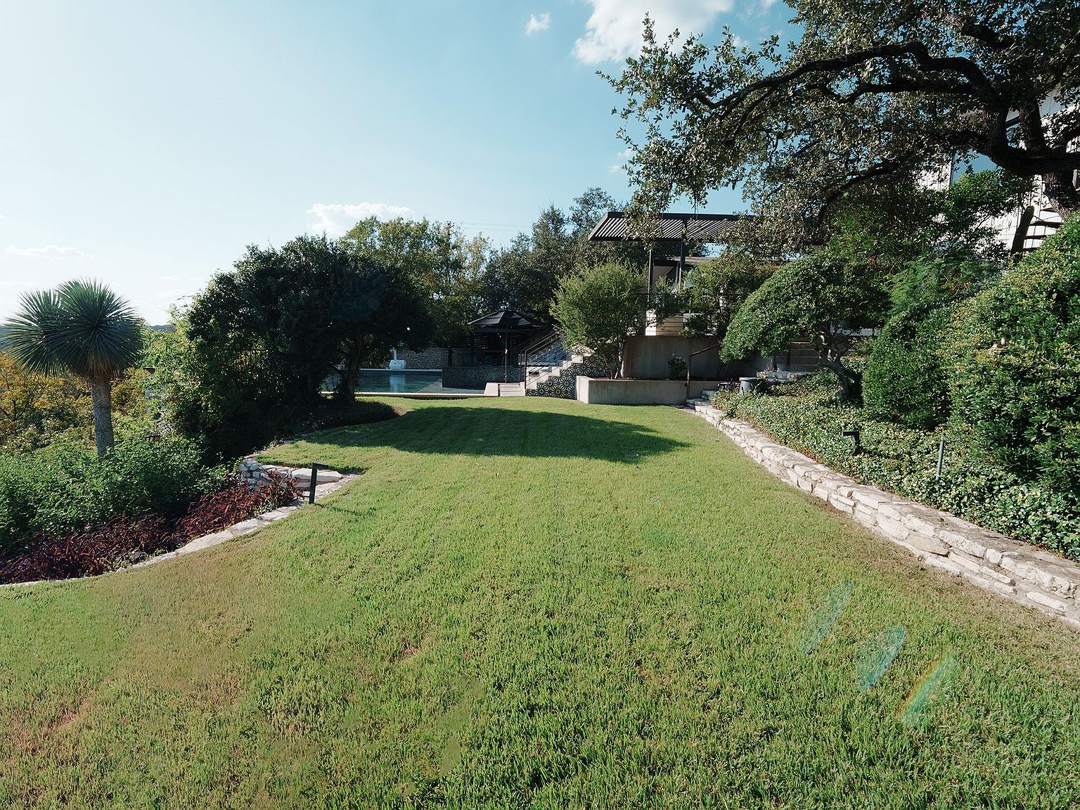 This image showcases a meticulously maintained yard with lush green grass, mature trees, and stone retaining walls. A gazebo and a glimpse of a pool can be seen in the background, suggesting a luxurious outdoor living space. The perspective is from a slightly elevated position, providing a wide view of the property.