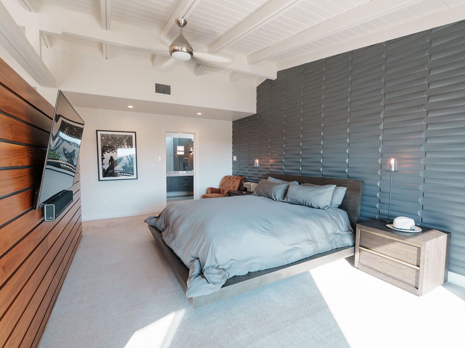 This is a primary bedroom featuring a modern design with a textured gray accent wall behind the bed. The room includes a large bed with gray bedding, a wooden nightstand, and a wall-mounted television on a wooden panel. The overall impression is clean, contemporary, and luxurious.