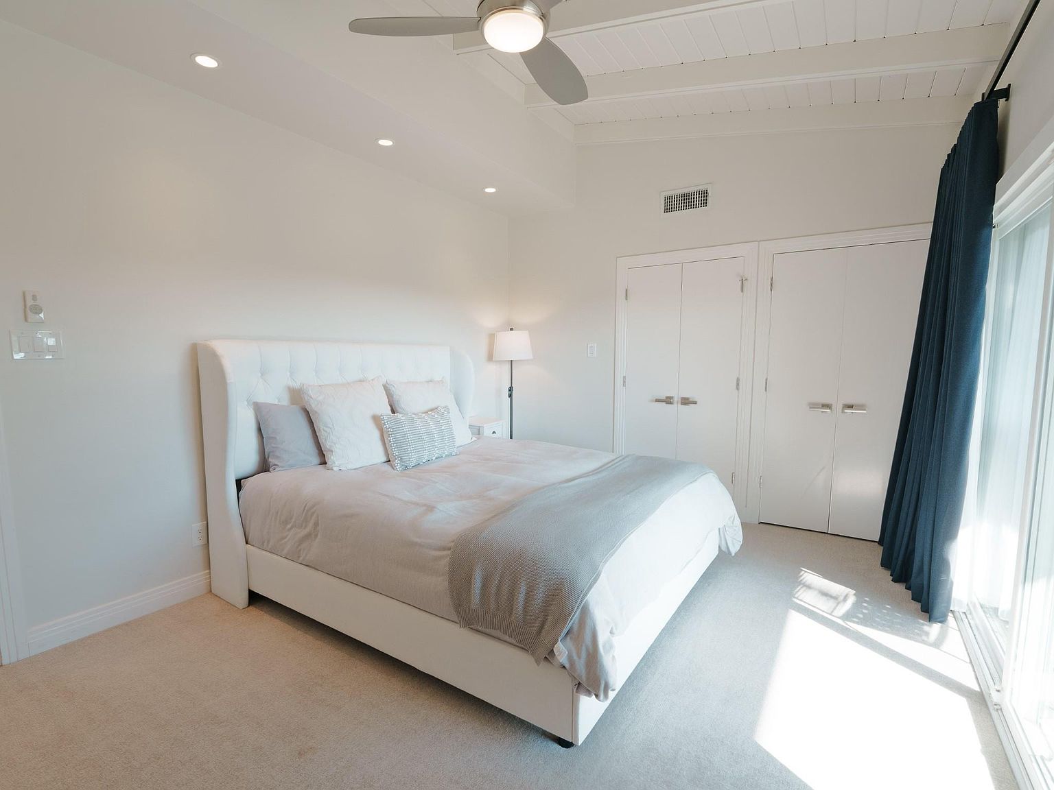 This is a bright and airy primary bedroom featuring a large white upholstered bed with neutral bedding and decorative pillows. The room has light-colored walls, carpeted floors, and recessed lighting, creating a serene and inviting atmosphere. Natural light streams in from a window with blue curtains, and there are white closet doors along one wall.