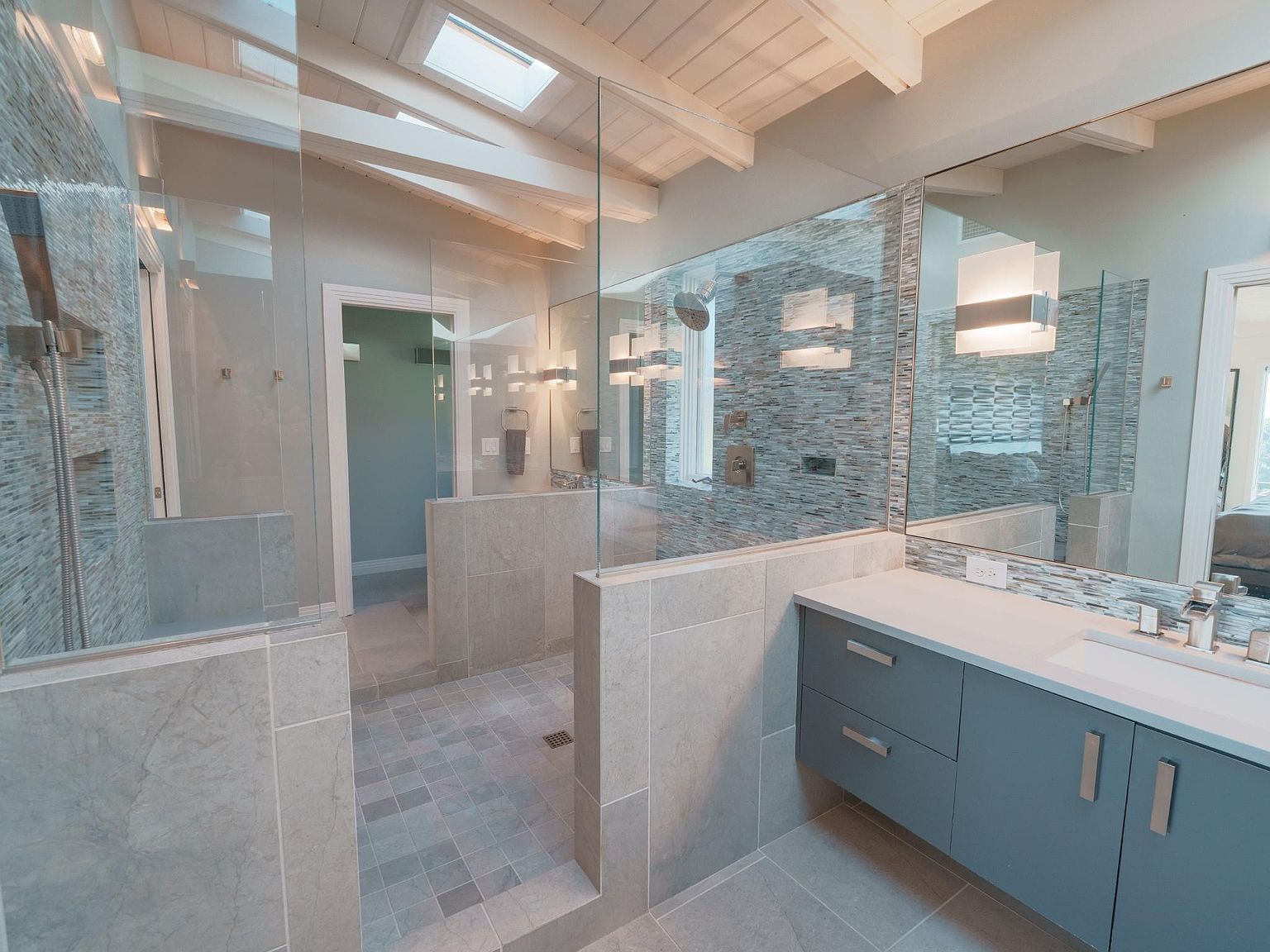 This is a modern primary bathroom featuring a glass-enclosed shower with mosaic tile accents and a rainfall showerhead. A large mirror reflects the vanity area, which includes a sleek, gray cabinet with a white countertop and modern fixtures. The bathroom has a clean and contemporary design with neutral tones and ample natural light from a skylight.