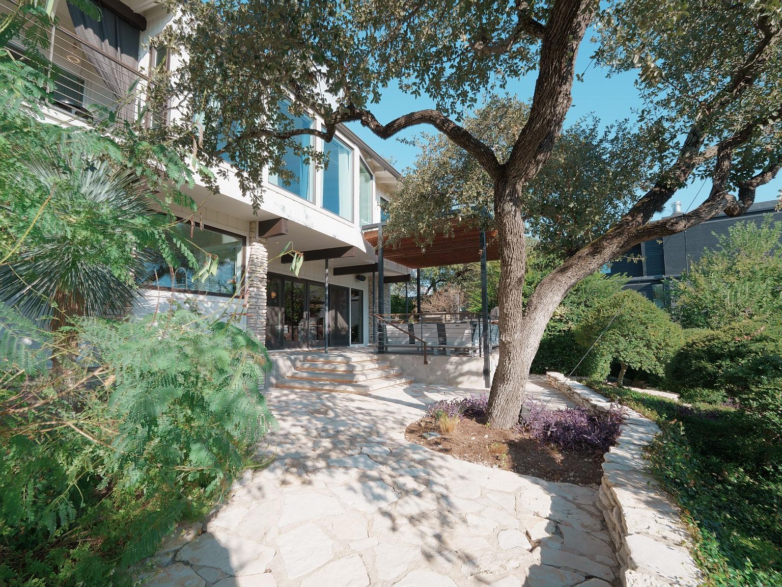 This image showcases a beautifully landscaped backyard featuring a stone patio and walkway. A large, mature tree provides shade and adds character to the space. The architecture of the house is visible in the background, blending seamlessly with the natural surroundings, creating an inviting outdoor living area.