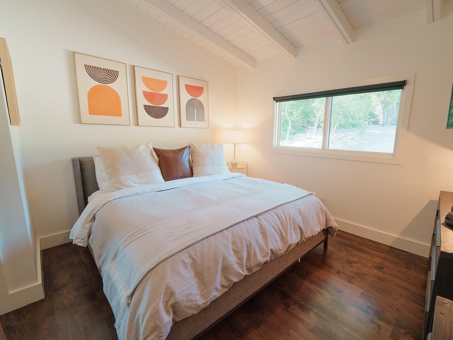 This is a well-lit primary bedroom featuring a queen-sized bed with neutral bedding and decorative pillows. Three framed art pieces hang above the bed, adding a touch of modern style. A window provides natural light, and the dark wood flooring complements the room's neutral color palette, creating a cozy and inviting atmosphere.