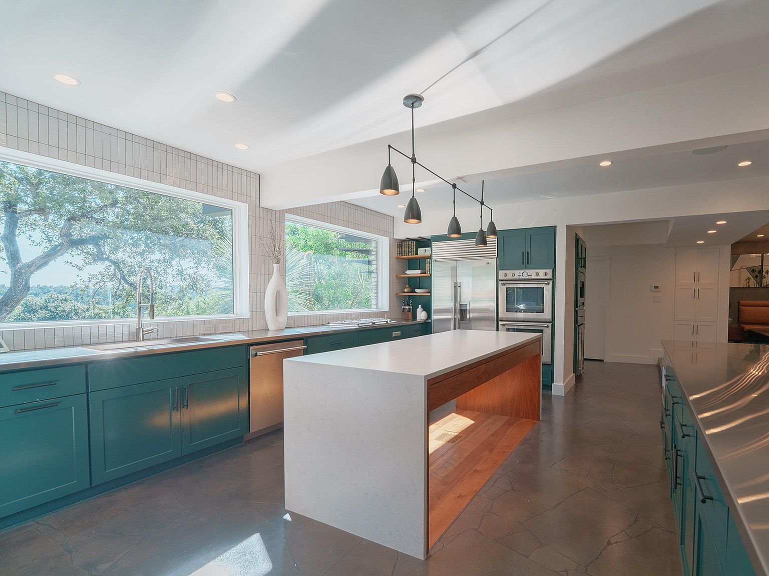 This is a bright and modern kitchen featuring teal cabinetry, stainless steel appliances, and a large island with a white countertop and wood base. Large windows provide ample natural light and views of the surrounding greenery. The kitchen also features recessed lighting and pendant lights over the island, creating a warm and inviting atmosphere.