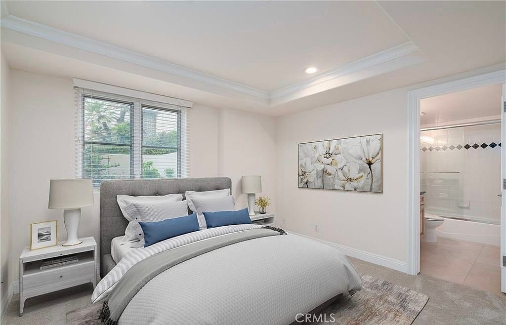 This bright and neutral bedroom features a queen-sized bed with a grey upholstered headboard, accented by blue throw pillows and a textured grey duvet. A white nightstand with a lamp sits to the left, while a large floral-themed wall art piece hangs above the bed. To the right, an open doorway provides a clear view into an adjoining bathroom with a shower-tub combination.