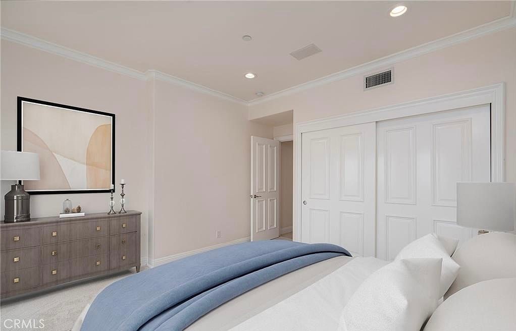 This bedroom features a neutral color palette with soft beige walls and white trim, creating a calm and inviting atmosphere. A grey dresser sits against the wall beneath a framed piece of abstract art, while a bed with a blue throw blanket occupies the foreground. The room includes white sliding closet doors and a doorway leading to an adjacent space, all illuminated by recessed ceiling lighting.