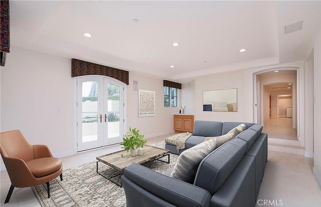 This bright and airy living room features a large, dark gray sectional sofa paired with a modern tan leather armchair. A textured area rug sits beneath a low-profile wooden coffee table, while a set of arched French doors with patterned valances provides access to the outdoors. The space is finished with neutral walls, recessed lighting, and a small built-in cabinet area in the corner, creating a comfortable and inviting atmosphere.