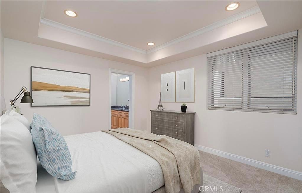 This bright and airy bedroom features a tray ceiling with recessed lighting and neutral-toned walls. A comfortable bed with patterned accent pillows is positioned in the foreground, while a wooden dresser sits against the wall beneath two framed art pieces. A doorway leads into an adjacent bathroom, and a large window with horizontal blinds allows natural light to fill the space.